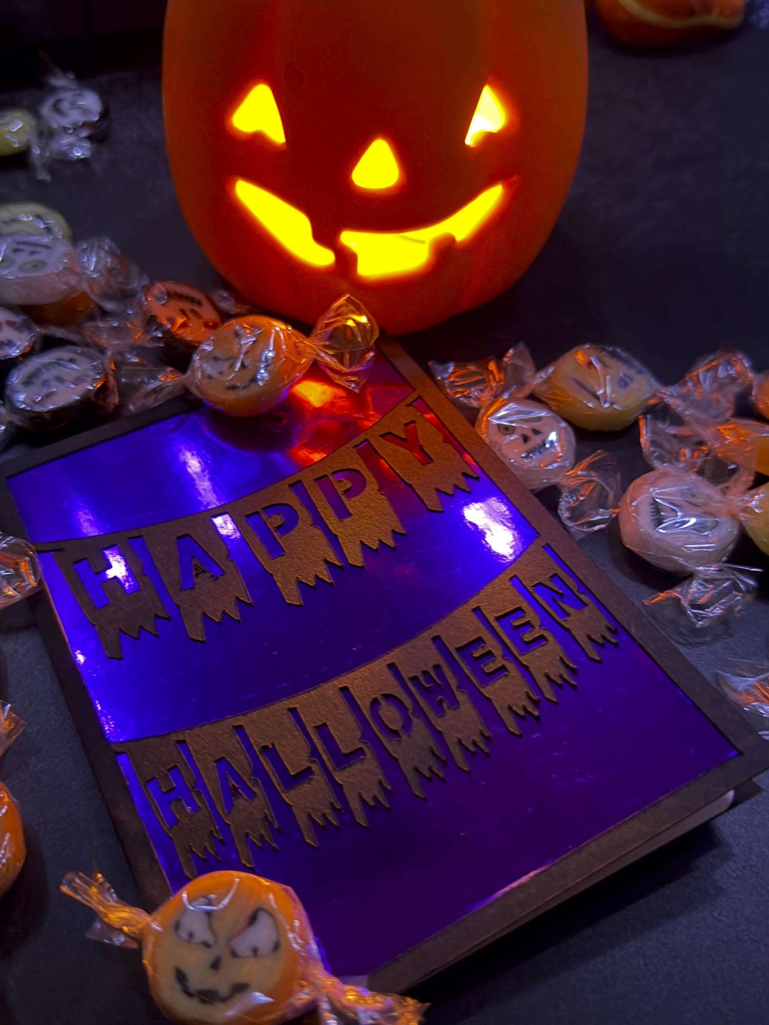 Geplottete Halloween-Klappkarte mit „Happy Halloween“-Wimpelketten und glänzend violettem Inlet, neben leuchtendem Kürbis und Bonbons.