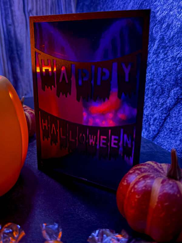 Geplottete Klappkarte „Happy Halloween“ mit zwei Wimpelketten und glänzendem Inlet, zwischen Kürbissen bei blau-violetter Beleuchtung.