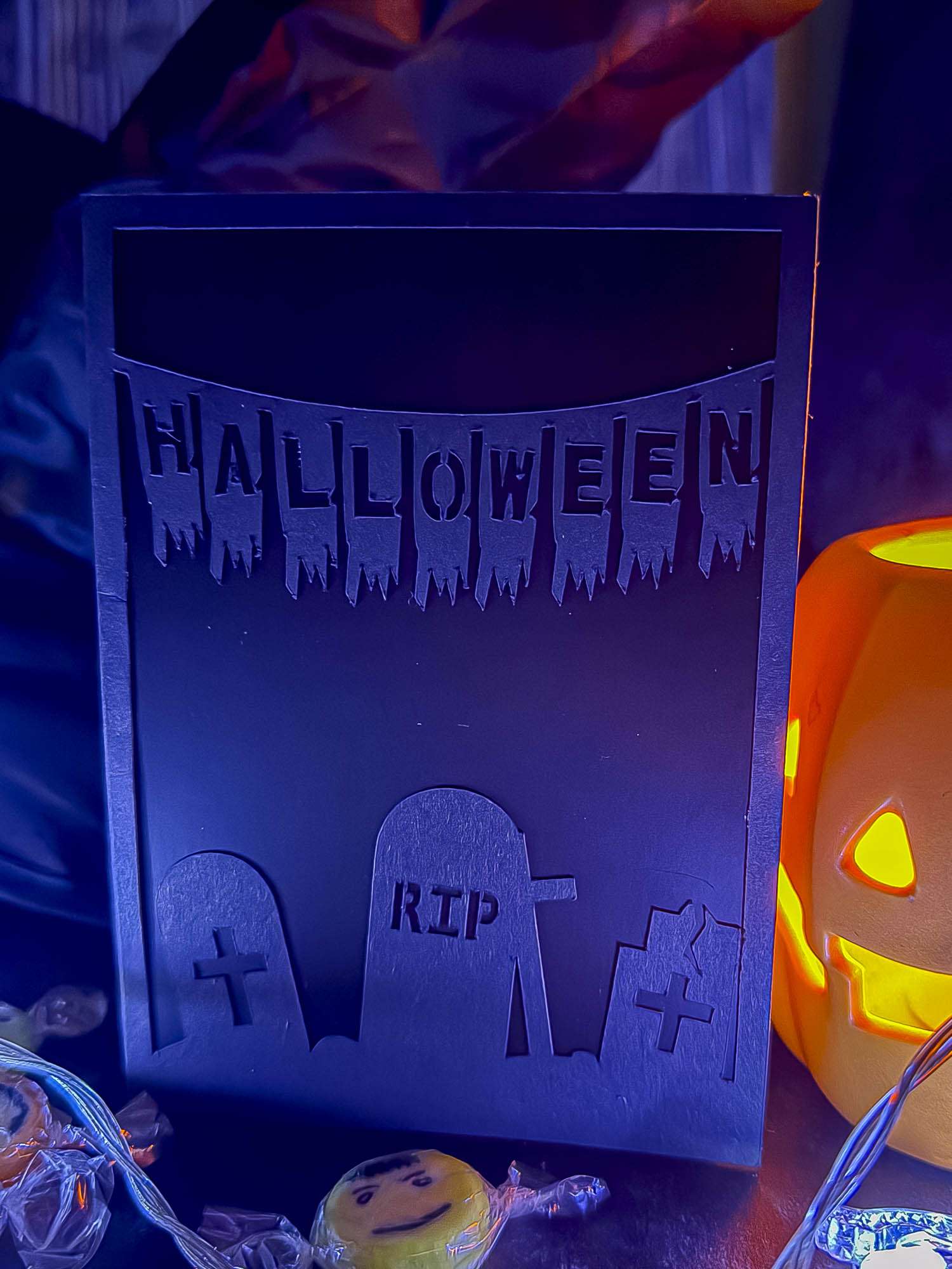 Geplottete Halloween-Klappkarte mit drei Grabsteinen und „Halloween“-Wimpelkette vor dunklem Inlet, neben einem leuchtenden Kürbis.