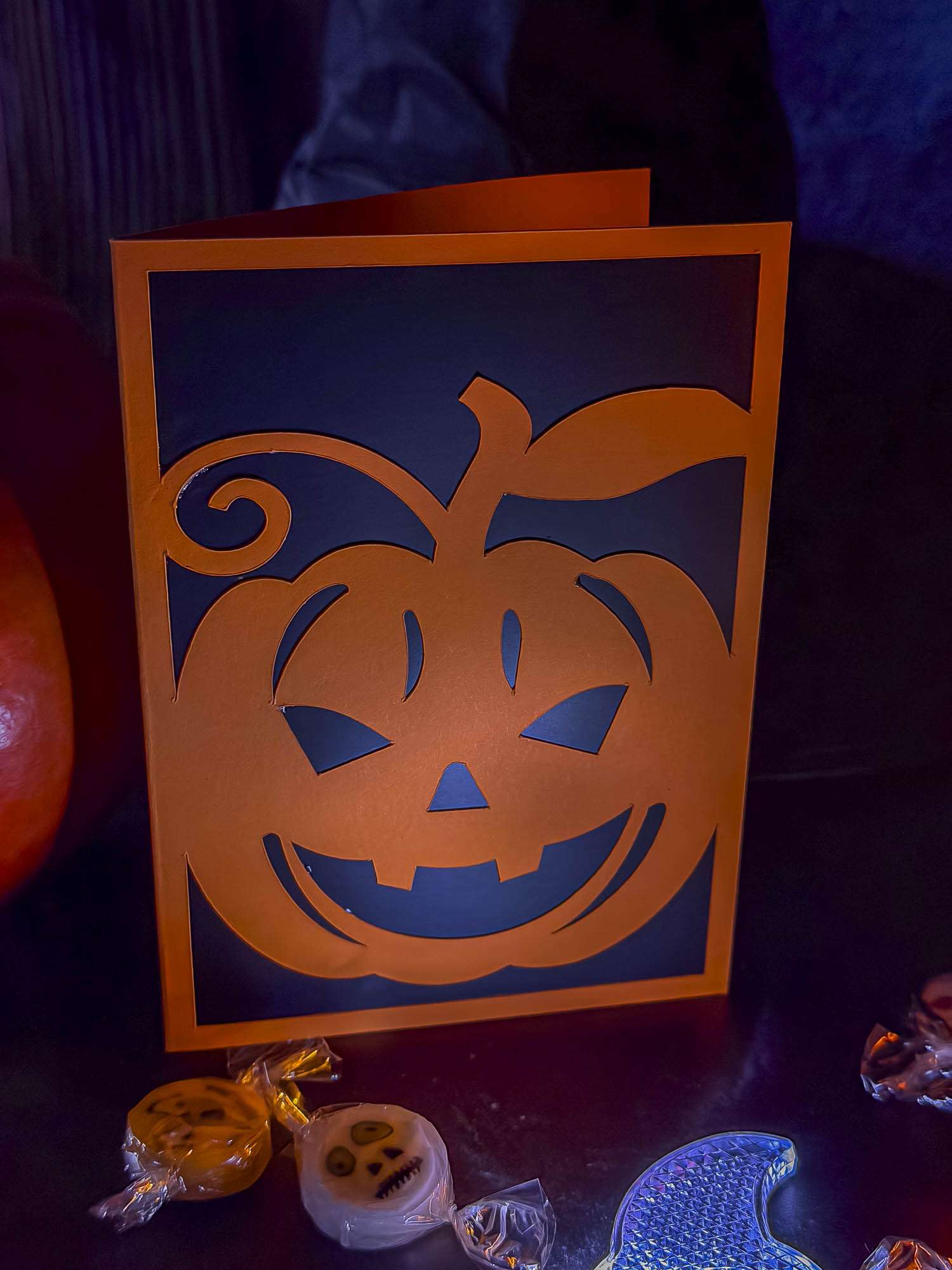 Orange Halloween-Klappkarte mit ausgeschnittenem Kürbisgesicht, Blatt und geschwungener Ranke vor dunklem Inlet.