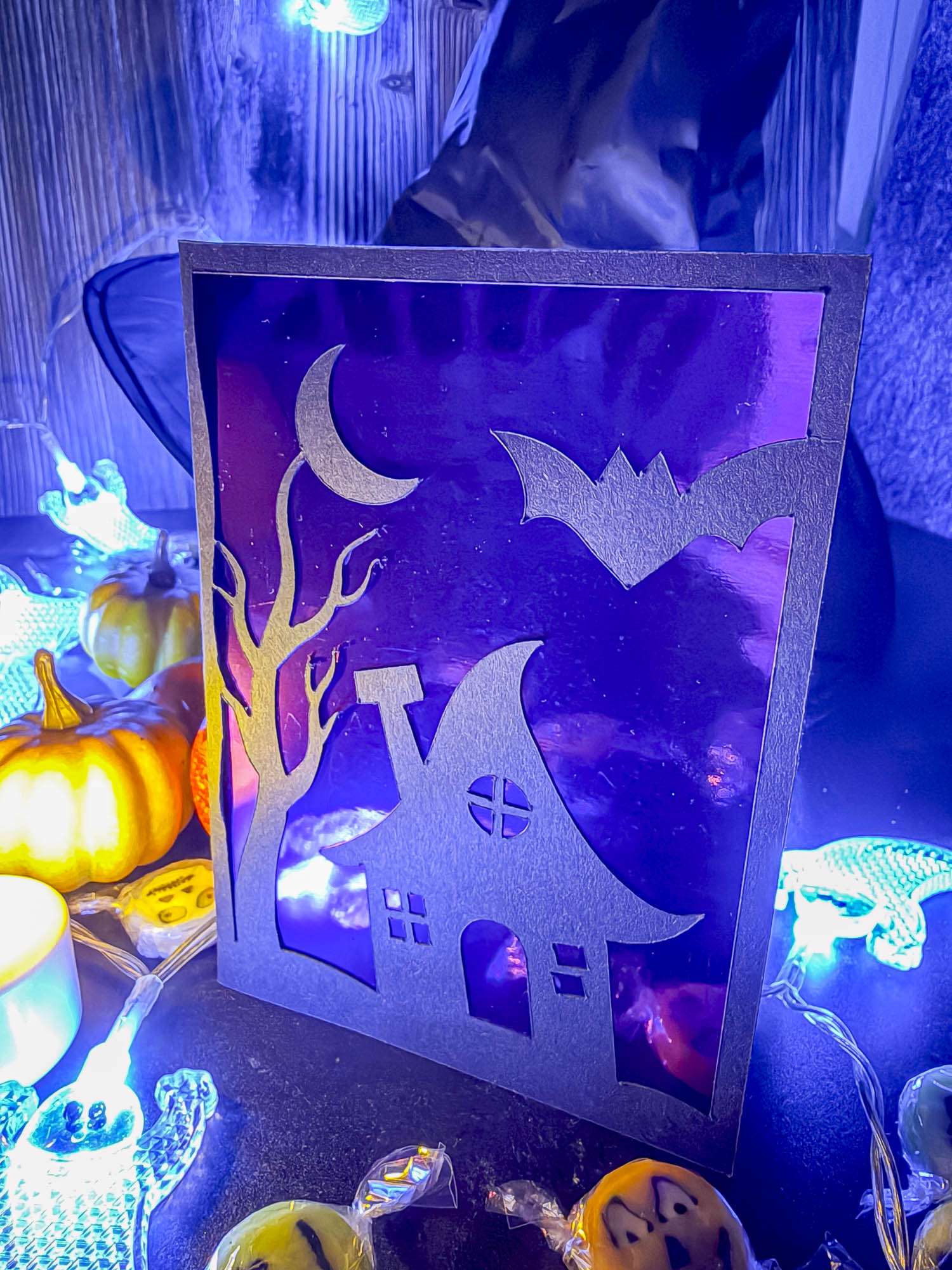 Geplottete Halloween-Klappkarte mit Hexenhaus, kahlem Baum, Mondsichel und Fledermaus vor glänzend violettem Inlet.