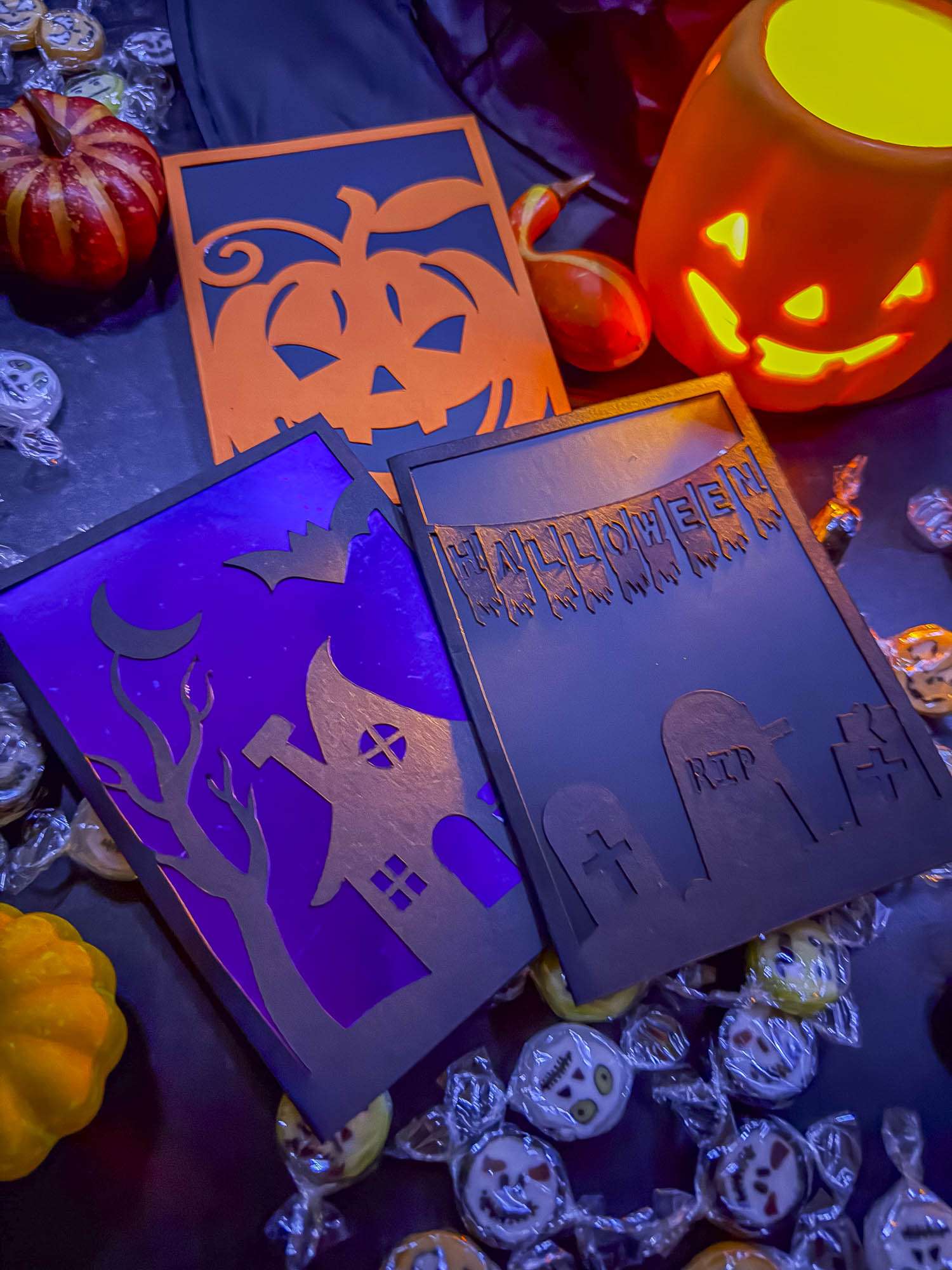 Drei geplottete Halloween-Klappkarten mit Kürbis, Hexenhaus und Friedhof, mit farbigen Inlets hinterlegt und zwischen Kürbissen und Bonbons arrangiert.
