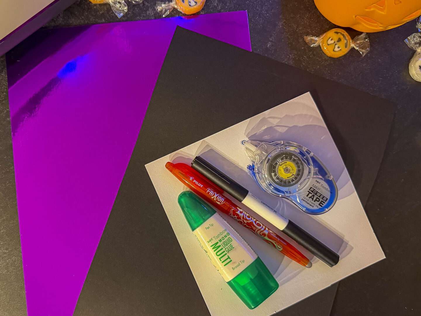 Material für eine Einladung mit Geheimtinte: schwarzer Tonkarton, violetter Effektkarton, weißes Papier, FriXion-Stift, schwarzer Zeichenstift, Flüssigkleber und Kleberoller.
